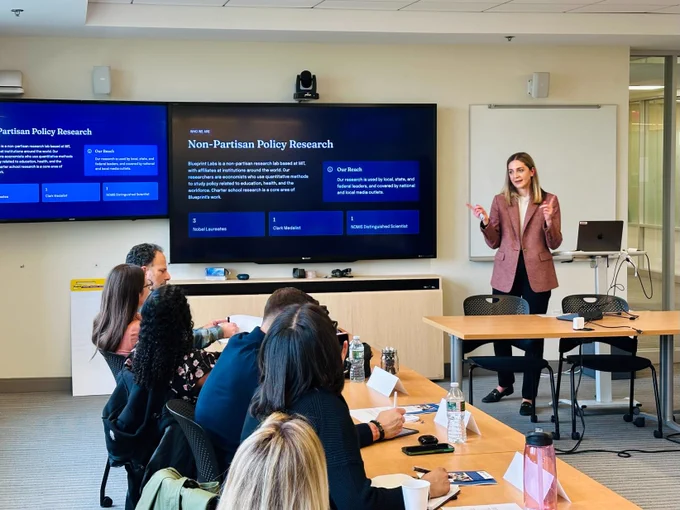 Blueprint Labs Executive Director Eryn Heying stands at the front of a classroom next to two screens showing a slide titled "Non-Partisan Policy Research." A row of charter authorizers sit at a table with and watch the presentation. 