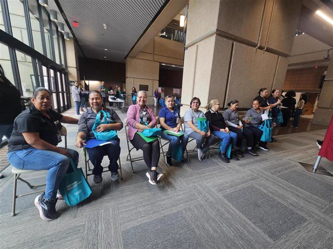 A group of people sitting in chairs along a hallway, holding teal-colored bags. The setting appears to be a building with large windows and carpeted floors.