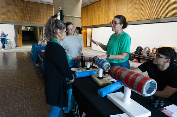 Individuals are engaged at a display table featuring several launchers made from colorful, taped tubes. The setting appears to be an indoor public area with various tables and informational materials. There are posters and banners visible in the background, and a few people are interacting with those at the table.