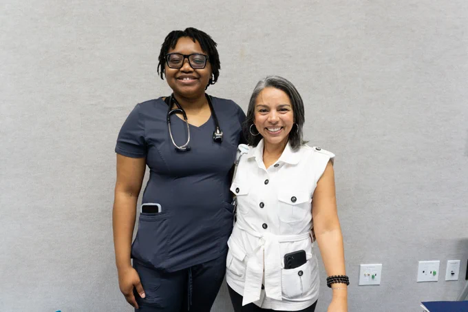 Two people are standing side by side, smiling. One is dressed in medical attire with a stethoscope around the neck, and the other is wearing a white, sleeveless outfit. 