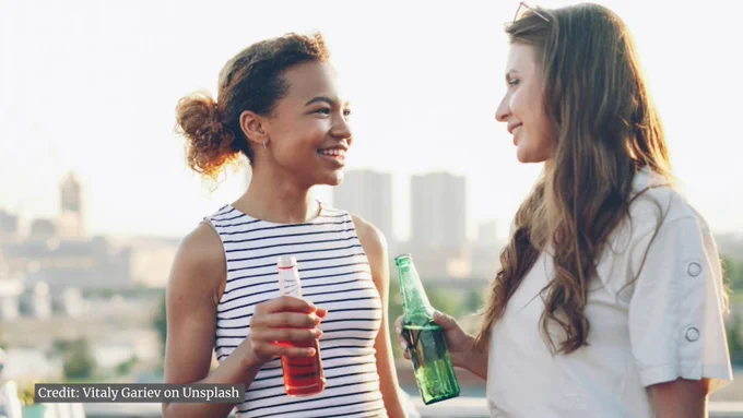 Two women standing outdoors in casual summer clothes, holding bottled drinks and chatting with a city skyline in the background.