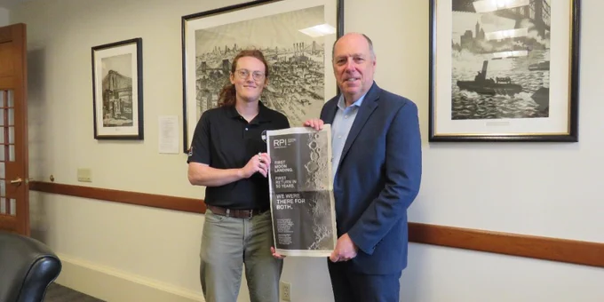 Two people stand in an office holding a copy of the New York Times, featuring an RPI print ad. They are surrounded by framed artwork on the walls.
