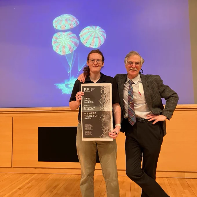 Two individuals stand on a stage, one holding a copy of the New York Times, featuring an RPI print ad. Behind them is an image of a space capsule with parachutes. They are both smiling, and the setting appears to be a seminar.