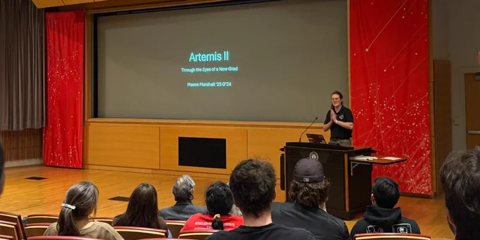 A person stands at a podium giving a presentation in a lecture hall. The screen displays the title "Artemis II: Through the Eyes of a New Grad." An audience sits facing the presenter.