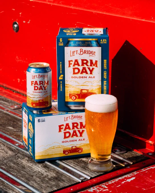 A rustic display of Lift Bridge Farm Day Golden Ale shows a six-pack, single can, and a freshly poured glass on a wooden surface against a bright red backdrop. The scene evokes a bright and refreshing ambiance.