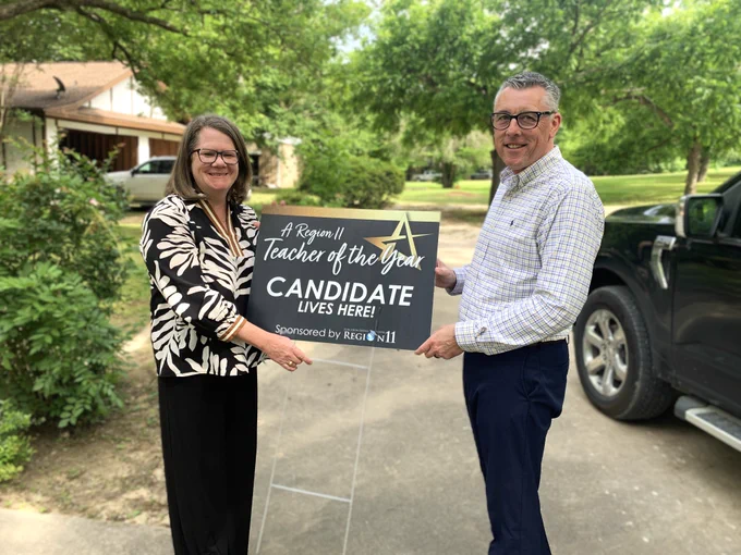Karen Allen and Kirby Basham stand outdoors, smiling, each holding the edge of a sign that reads, "Region 11 Teacher of the Year Candidate Lives Here!" A truck is visible in the driveway, with trees and a house in the background.