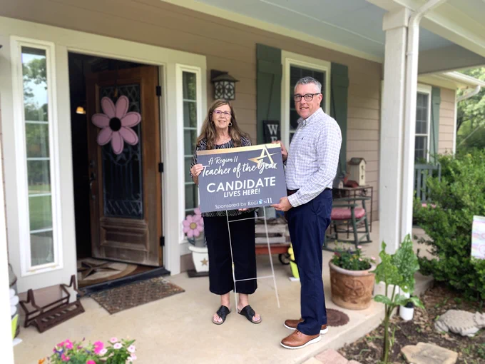 Laurie Anthony and Kirby Basham stand on a porch holding a sign that reads "A Region 11 Teacher of the Year Candidate Lives Here!" The house has a decorated entrance with flowers and a potted plant.