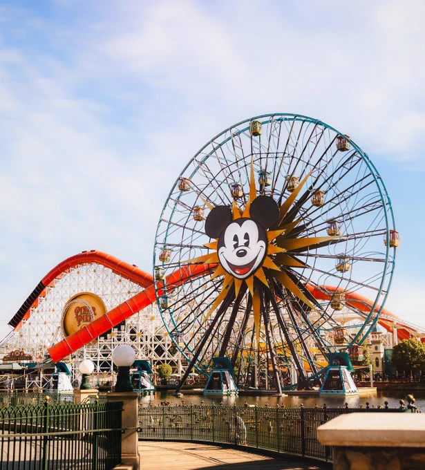 A large Ferris wheel with Mickey Mouse's face at the center stands near a colorful roller coaster. The scene is part of Pixar Pier at Disneyland, surrounded by a bright blue sky.