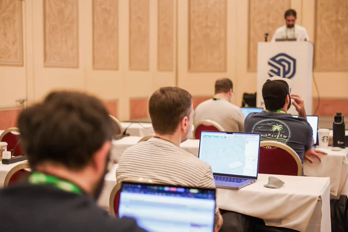 Conference attendees work on laptops while a speaker presents at a podium in a hotel meeting room.