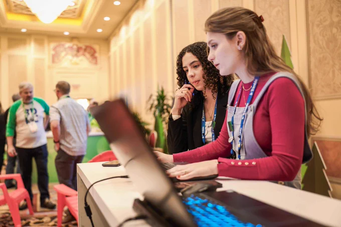 Two women wearing conference badges work at a laptop kiosk in a hotel ballroom while attendees mingle in the background.