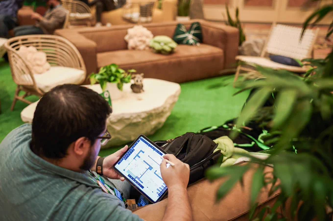 Man seated in a lounge uses a tablet and stylus to view a floor plan, surrounded by plants, wicker chairs, and a brown sofa.