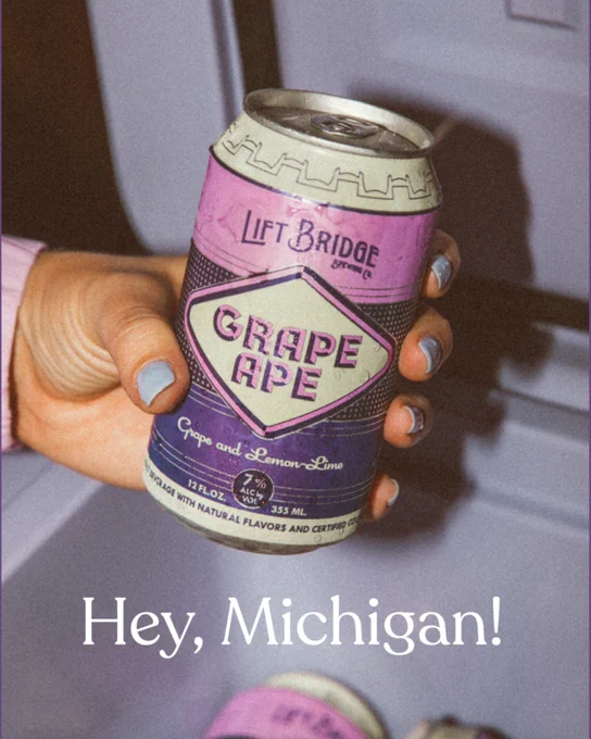 A hand holding a can of Lift Bridge's Grape Ape, featuring grape and lemon-lime flavors. The text overlay says 'Hey, Michigan!' with a casual and friendly tone.