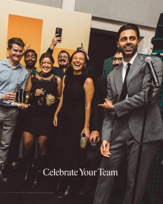 A group of colleagues joyfully celebrating with drinks and awards at a corporate event. An engaging speaker in a suit is pointing towards them.