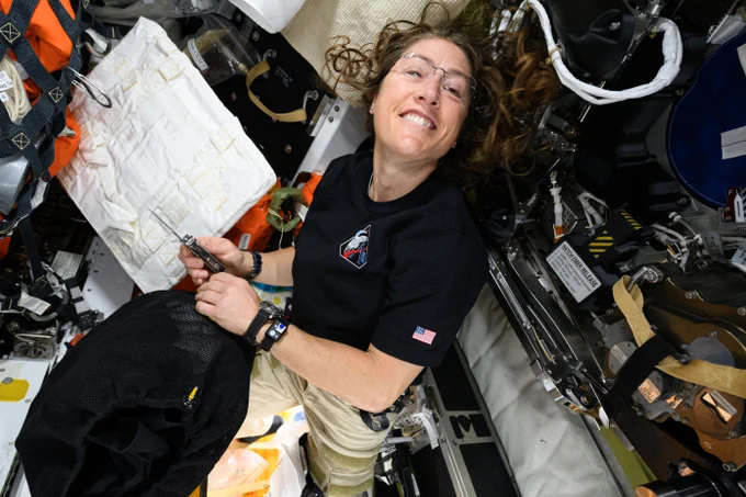 Christina Koch smiles at the camera as she holds a tool in one hand and a black mesh bag in the other. Credit: NASA