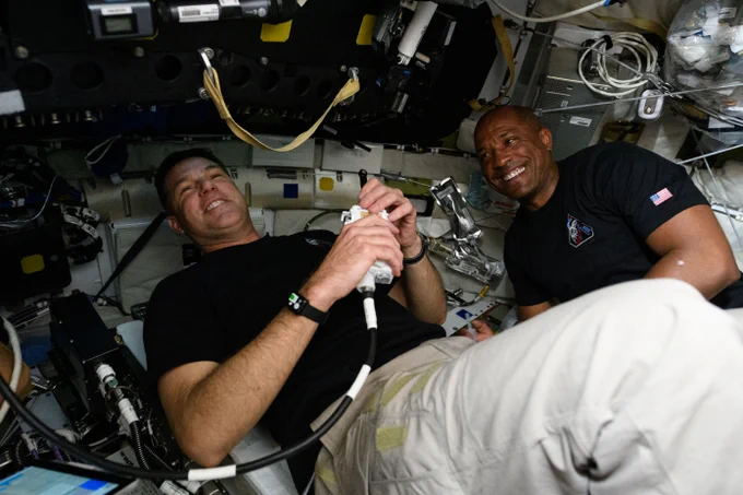 Hansen and Glover smile and laugh inside the spacecraft. Hansen can be seen holding a tool that is connected to a wire. Credit: NASA