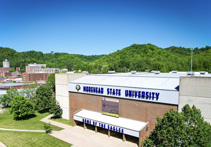 An aerial shot of Morehead State University’s campus with the main focus being on a large facility known as the Academic Athletic Center. The front of the building has large blue letters spelling out “Morehead State University” and “Home of the Eagles.” Several other university buildings are in the distance as well as the lush forested hills surrounding the the campus.