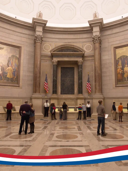 Grand room with large columns and 2 American flags in the National Archives with approximately 10 vistors looking at displays and 2 security guards