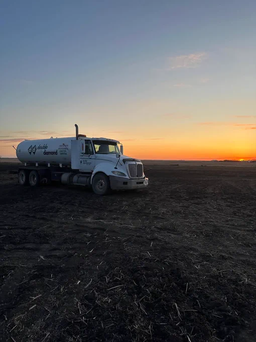 A NH3 fertilizer delivery truck with "Double Diamond" branding is parked on a dark, expansive field at sunset. The sky is a gradient of warm colors, and the horizon is clear.