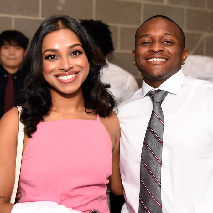 Two people smiling at the camera. They are wearing formal attire and there are people in the background.