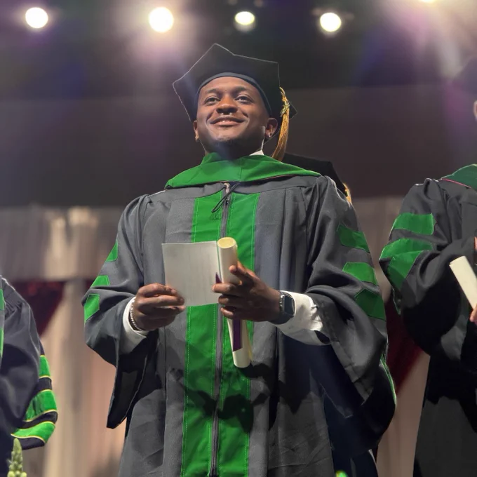 A person in a graduation cap and gown is smiling and holding a diploma on stage. There are bright lights overhead and others in similar attire nearby.