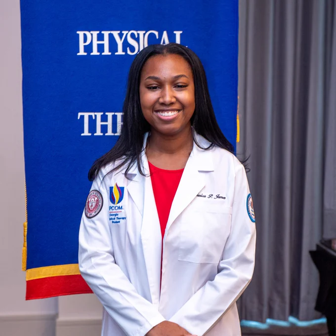 A person wearing a white coat is smiling in front of a blue banner with the words "Physical Therapy" partially visible. The coat features patches with the Philadelphia College of Osteopathic Medicine (PCOM Georgia) insignia.