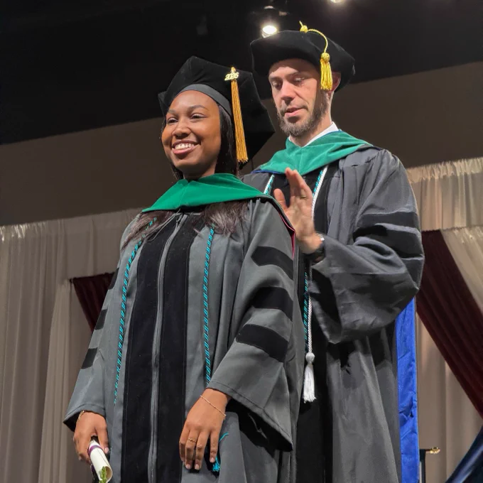 A person in graduation attire stands smiling on stage, while another in similar attire adjusts their hood. Both are in front of a curtain backdrop.