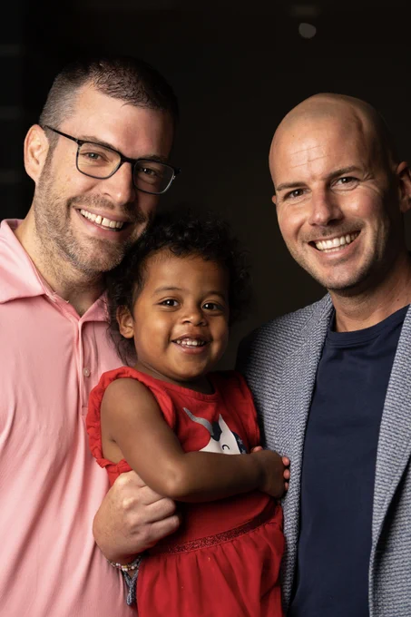 A person in a pink shirt and glasses stands next to a person in a blazer, both smiling at the camera. They are holding a young child wearing a red outfit with a Minnie Mouse design, who is also smiling.