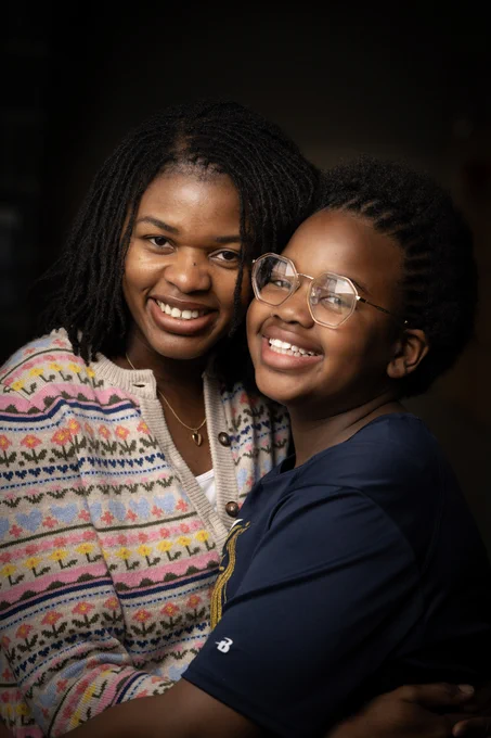 Two people are smiling and embracing. One is wearing a colorful patterned sweater, while the other wears glasses and a dark shirt.