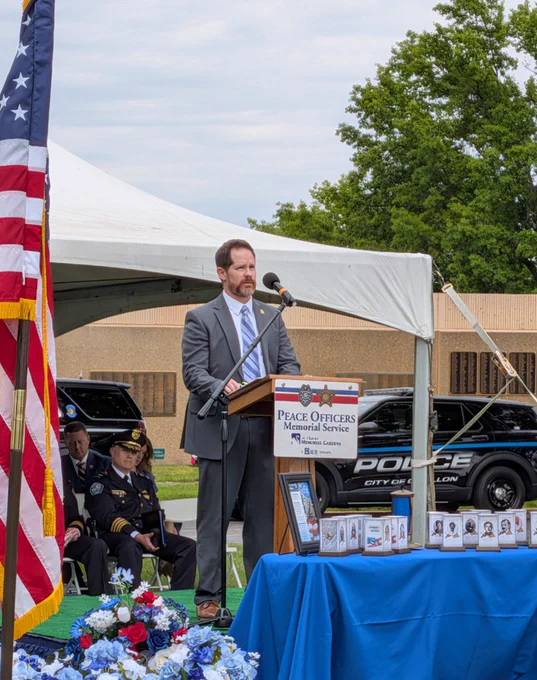 Man in suit speaking at podium during outdoor Peace Officers Memorial Service with police officers seated nearby.