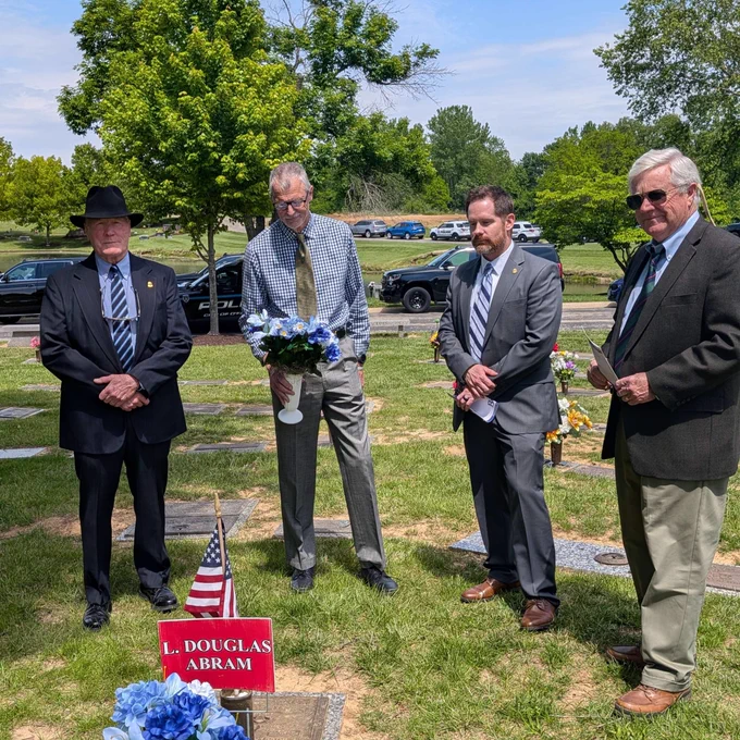 Four men in formal attire standing solemnly around a gravesite, which is adorned with a U.S. flag and blue flowers.