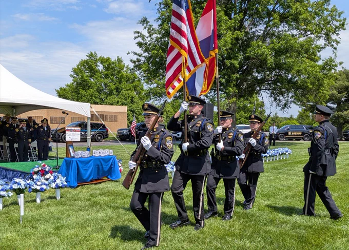 Uniformed police officers march in formation carrying U.S. and Missouri flags during an outdoor ceremony on a sunny day.