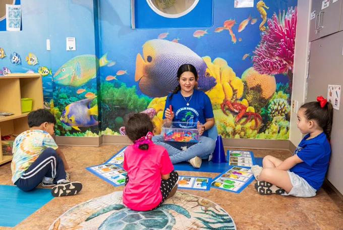 A group of children sits on the floor around a TAMU-CC student teacher holding a toy bin in a colorful, sea-themed room with vibrant images of fish and coral on the wall. Educational cards are spread out on the floor.