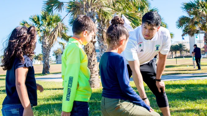 A group of young people and a TAMU-CC student teacher interact in a park with palm trees. The adult wears a white shirt with Texas A&M University-Corpus Christi branding visible on one person's sleeve. Buildings and more people are visible in the background.