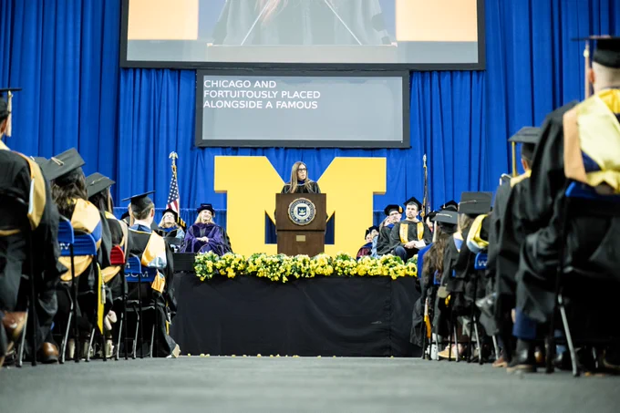 Jenny Just, BBA '90, speaking at a lectern on a stage decorated with a large Block M logo. The audience is filled with individuals in graduation attire. 
