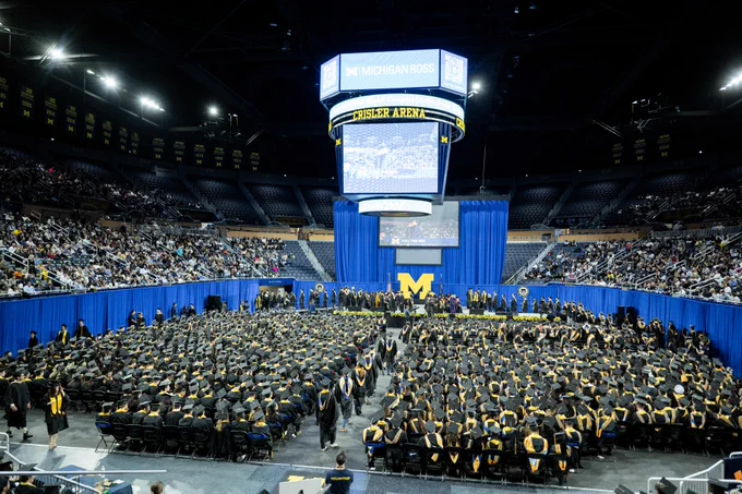 Large indoor graduation ceremony at Crisler Arena. Graduates in caps and gowns are seated in rows facing a stage. A big screen displays "Michigan Ross." The arena is filled with spectators.