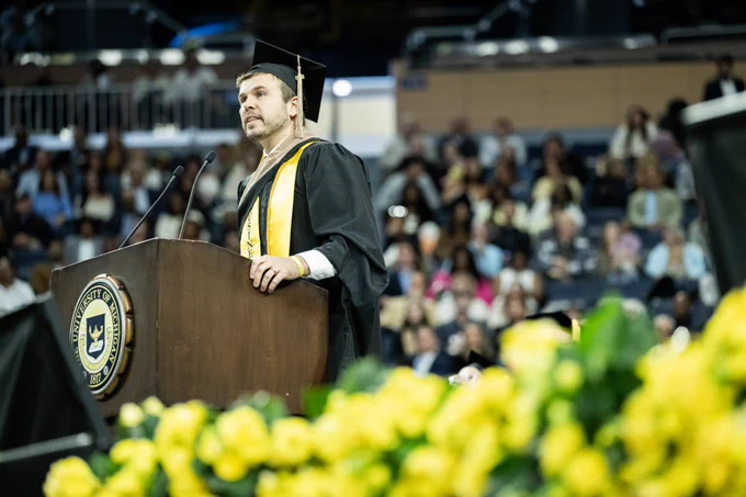 A person in a graduation cap and gown speaks at a podium with the University of Michigan emblem. The background is filled with an audience seated in the Crisler Center, and yellow flowers are visible in the foreground.
