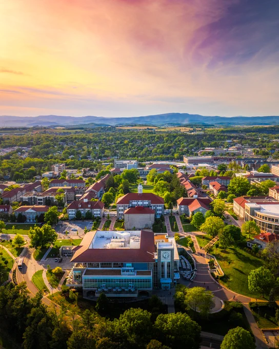 A sunset with yellow, pink and purple hues fill the sky above the Blue Ridge Mountains. In the foreground is the West Campus of JMU with Wilson Hall in the center.