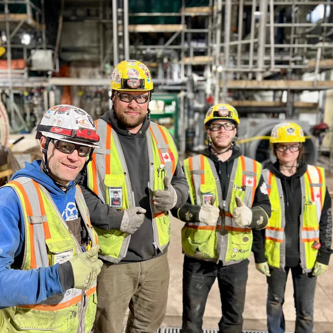 A group of four individuals wearing safety gear, including helmets with stickers and reflective vests, stand in an industrial setting. They are giving thumbs up and smiling. The background shows machinery and scaffolding. One of the helmets features a logo that includes "Canada.