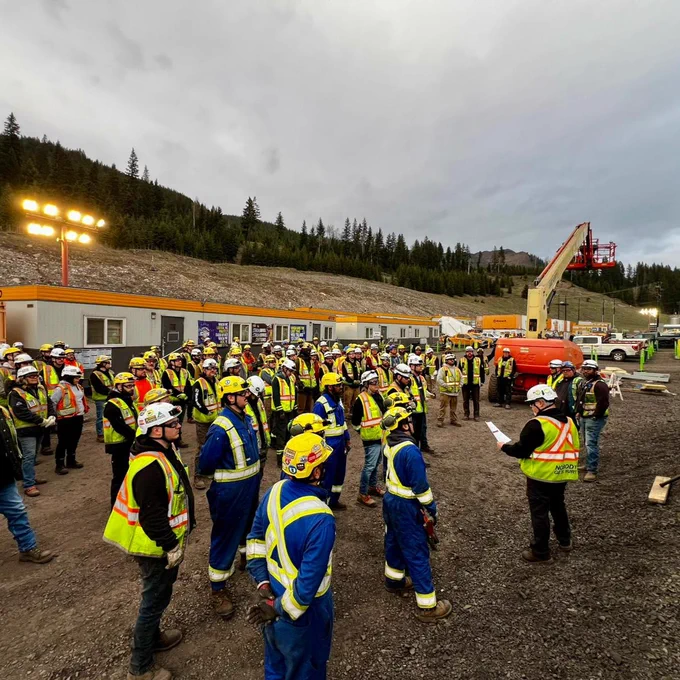 A group of construction workers wearing safety gear gathered outdoors on a worksite. The site includes portable buildings, a cement silo, and construction equipment. Trees and hills are visible in the background.