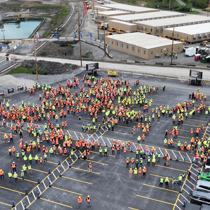Aerial view of a large group of people wearing high-visibility vests gathered in a parking lot. Nearby are several rows of parked vehicles and temporary office trailers. The scene appears to be at a construction site.