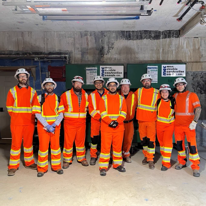 A group of people wearing orange safety uniforms and helmets stand together in a construction setting. Some are wearing reflective vests. Behind them is construction equipment and a wall with a signs. 