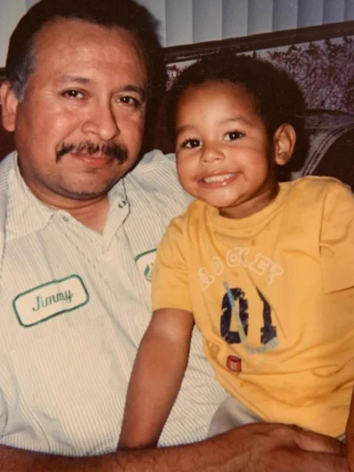 A man and a young child sit together, smiling warmly at the camera. The man is wearing a work shirt with a name tag, and the child is wearing a yellow t-shirt.