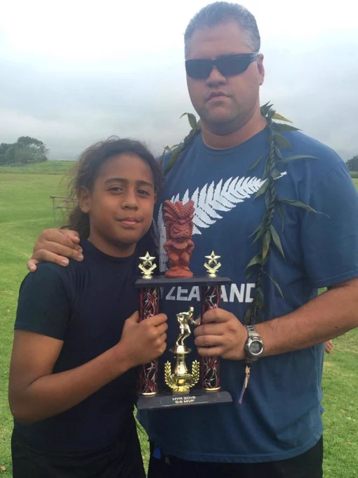 A father and son proudly display a sports trophy outdoors. The son holds the trophy as the father stands beside him, wearing a New Zealand-themed shirt and lei.
