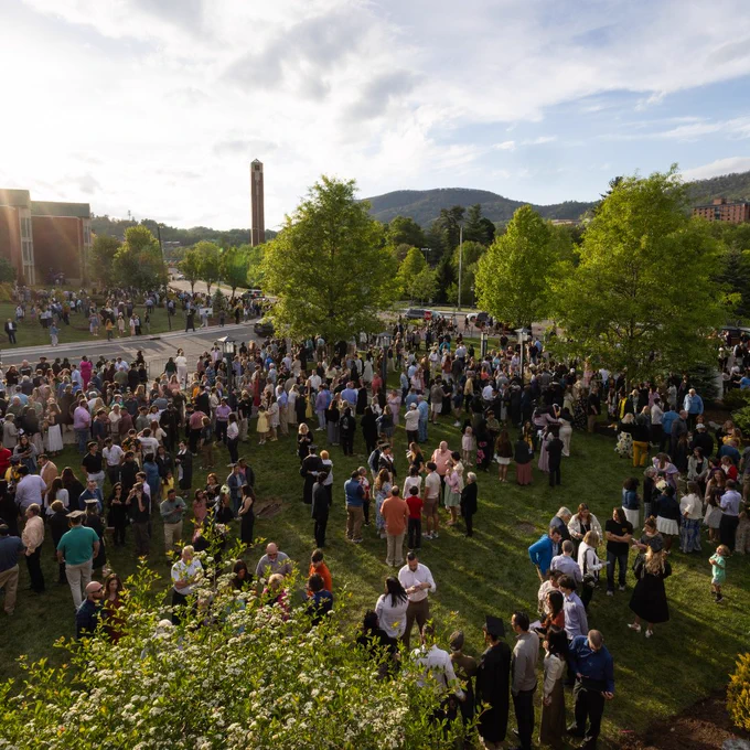 A large crowd gathers on a grassy area outside a building, with trees and distant hills in the background. A tall clock tower and more buildings can be seen under a partly cloudy sky.