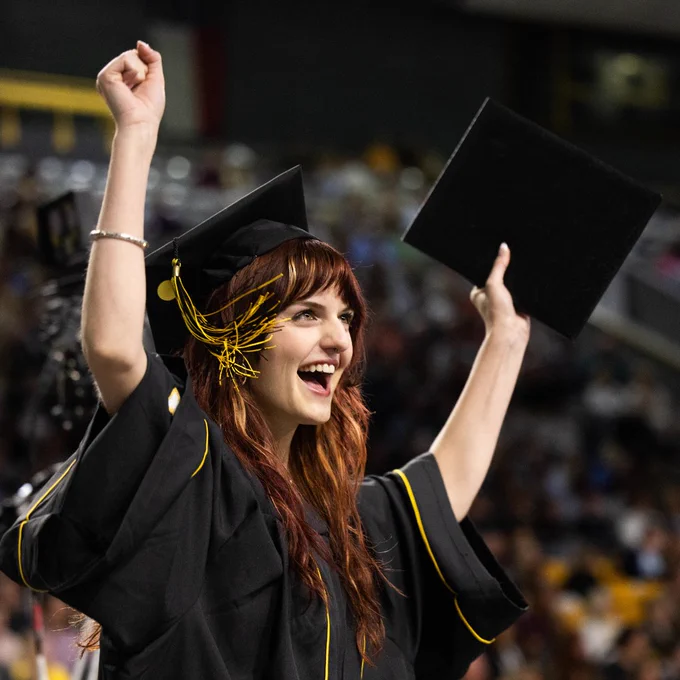 A graduate in a black cap and gown celebrates with a diploma in one hand. The person is smiling and raising both arms in a crowded ceremony setting.