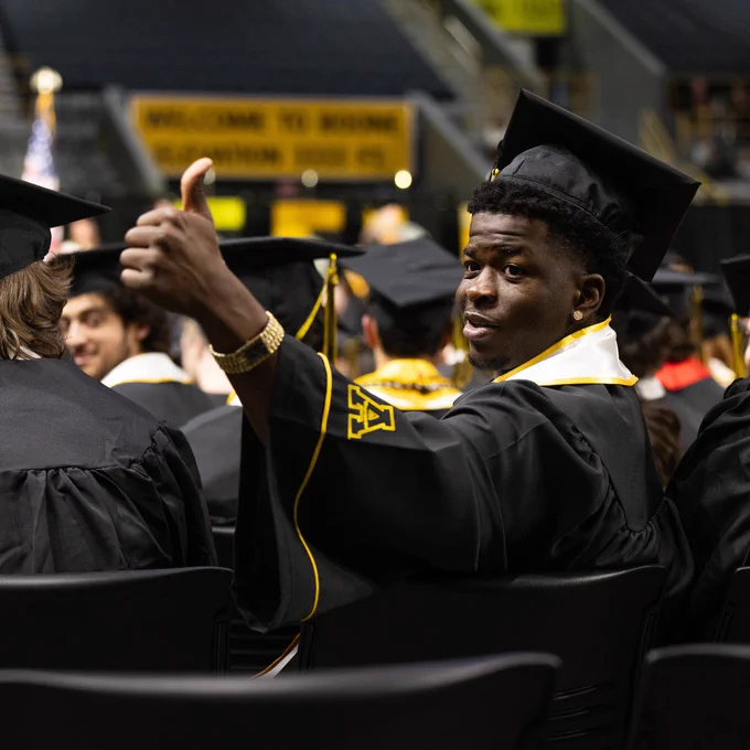 A graduate in a cap and gown gives a thumbs-up during a commencement ceremony. The background features a partially visible banner with the words "Welcome to Boone”. 