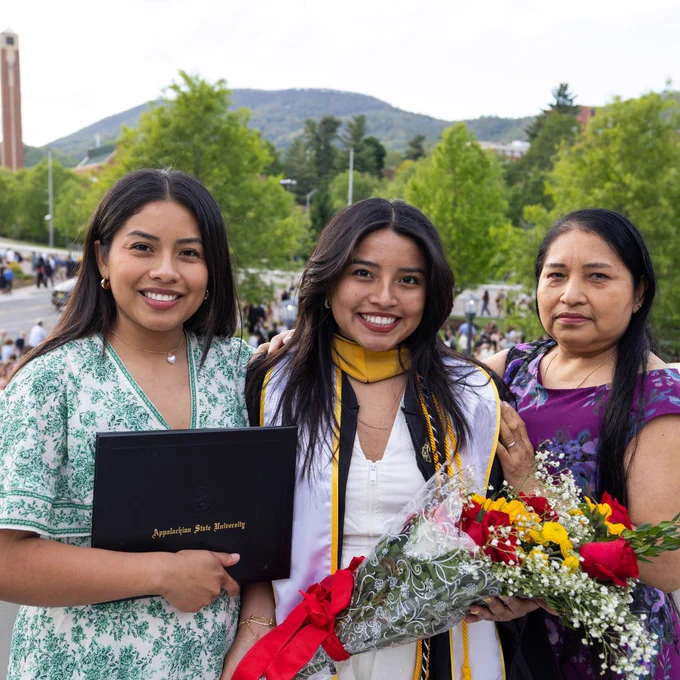 Three people are smiling outdoors in a celebratory setting. The center individual is wearing a graduation gown and holding a diploma from Appalachian State University. They are accompanied by two others, one holding a bouquet of flowers. Behind them, there is a scenic view of trees and hills.