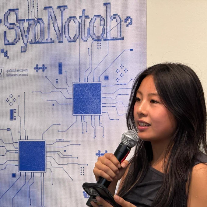 A person speaking into a microphone stands in front of a display with blue circuit patterns and the text "SynNotch".