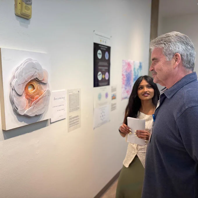 A gallery wall featuring scientific artwork from the HEAL Program at Keck School of Medicine of USC, with a researcher and artist standing in conversation.