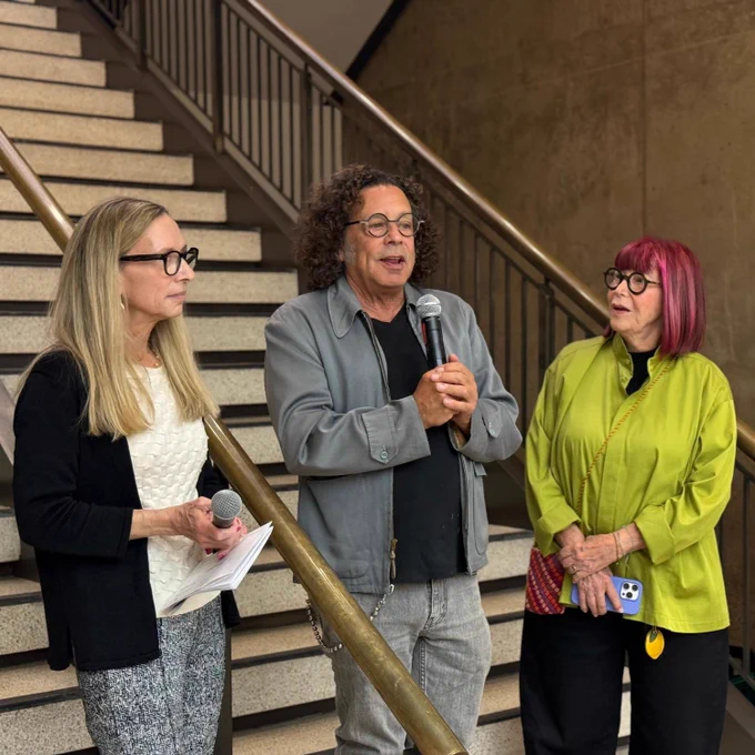 Three people stand in conversation on a staircase. One person is holding a microphone, while the others listen attentively. They are casually dressed, and the setting appears to be informal.